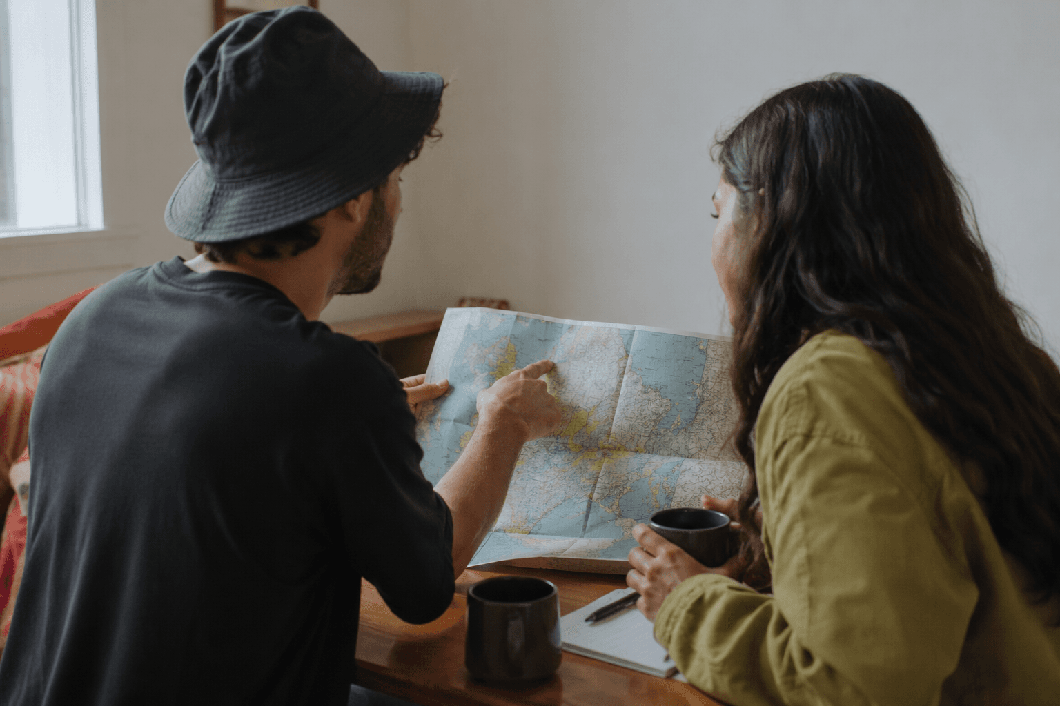 Two travellers at a table with an open paper map and coffee between them.
