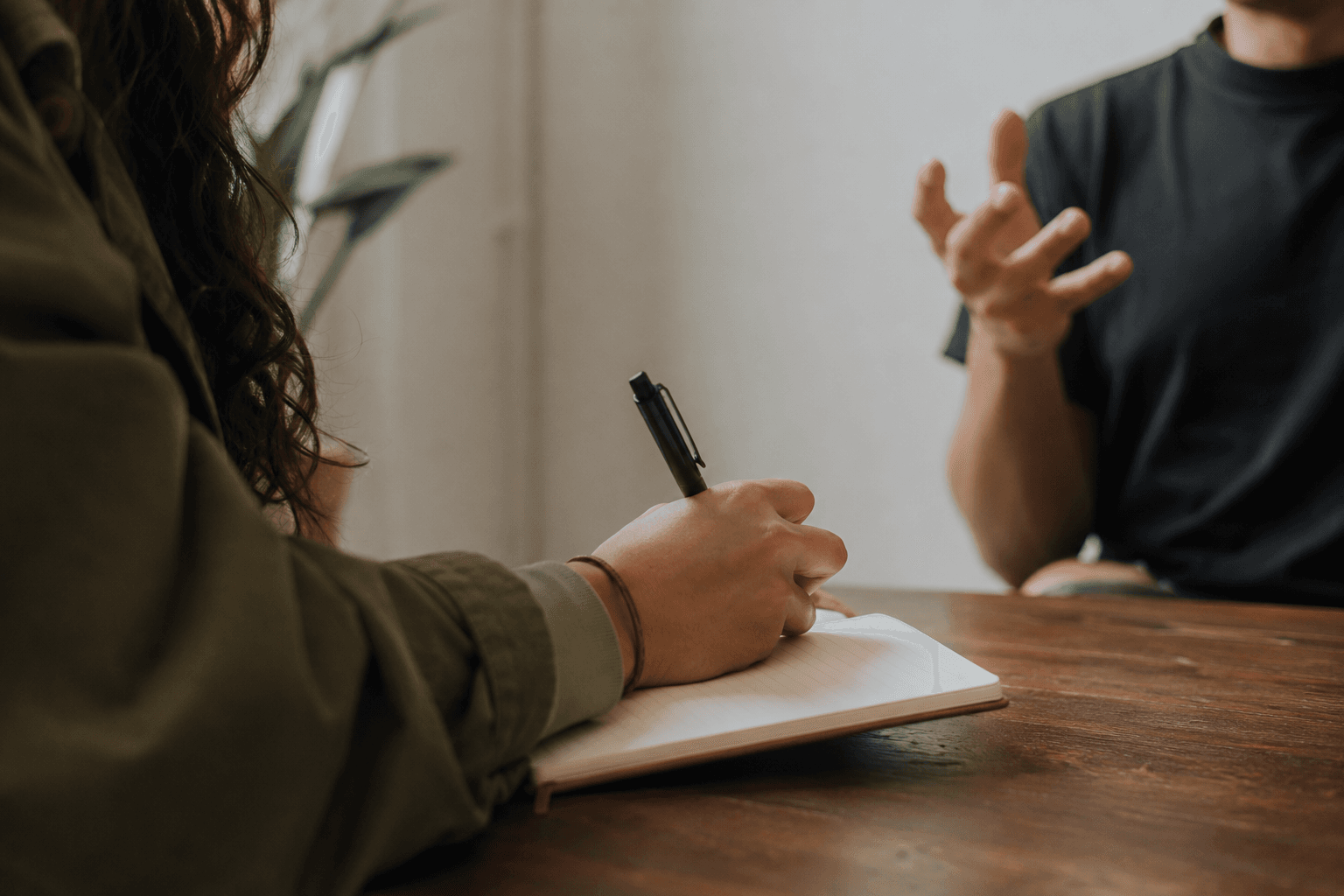 A hand writing in a notebook while someone across the table talks with their hands.