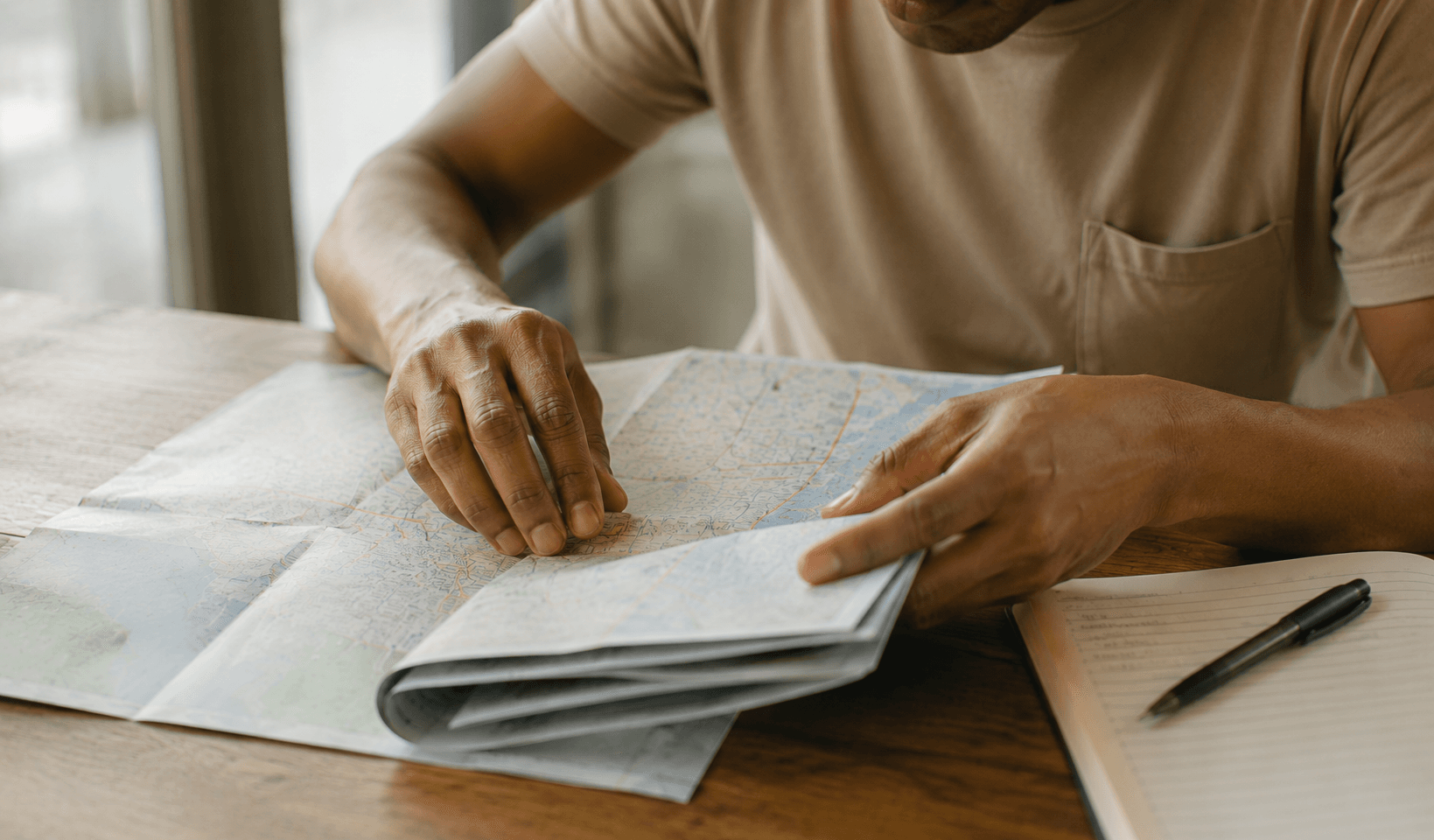 Hands smoothing a folded map flat on a table next to an open notebook.