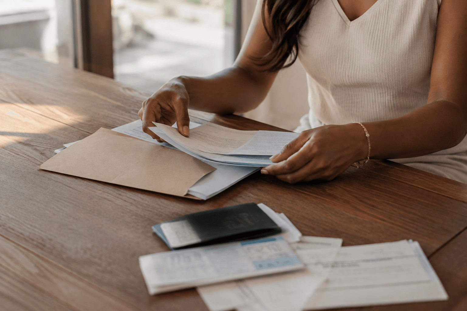 A woman sorting through documents and envelopes at a sunlit table.