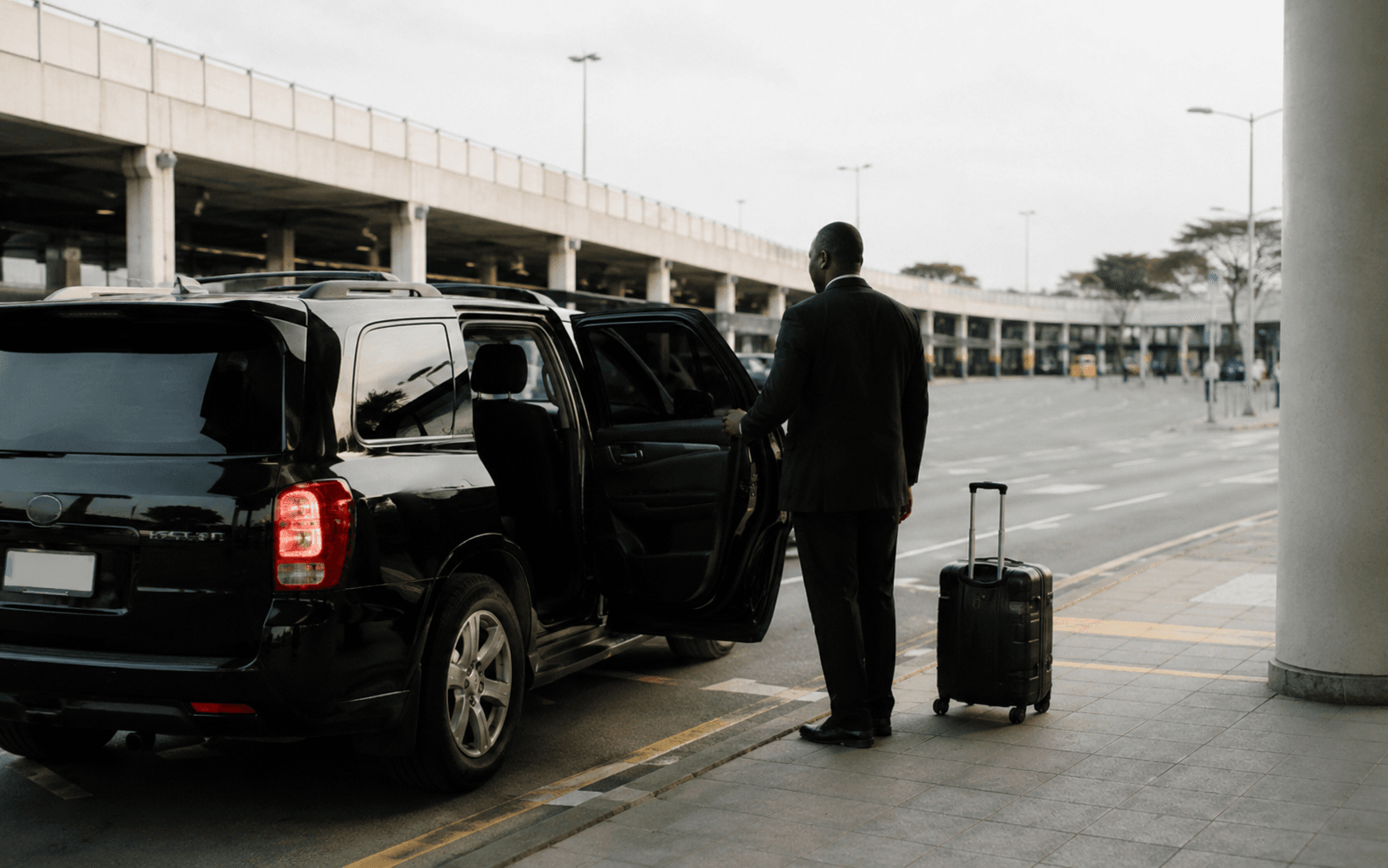 A driver holding open the rear door of a dark car at an airport kerb, a suitcase beside it.